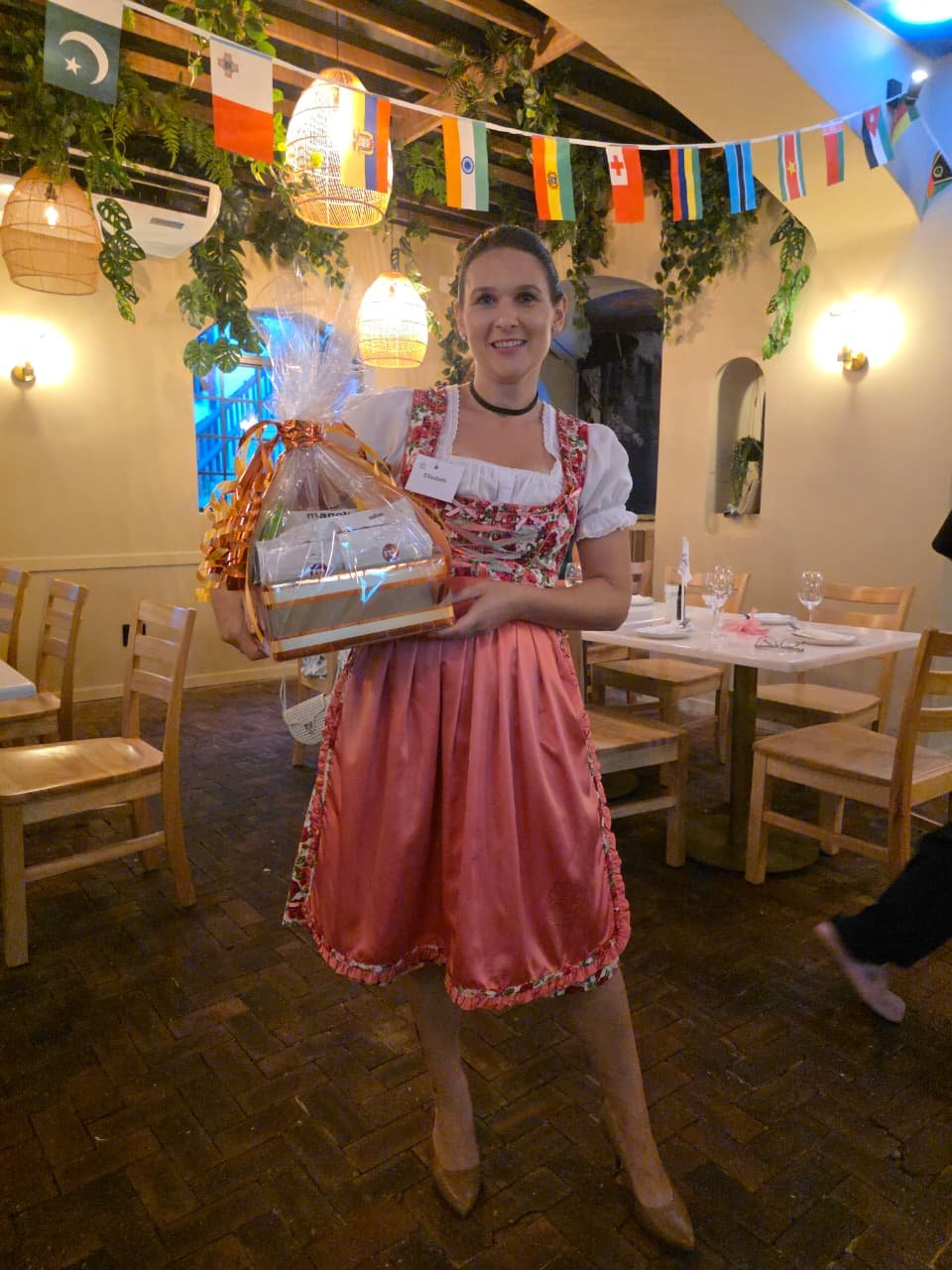 IWC Durban member in traditional dirndl holding a ribboned raffle prize basket beneath a canopy of international flags